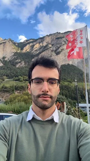 Ali Lari outdoors, wearing glasses and a green jumper